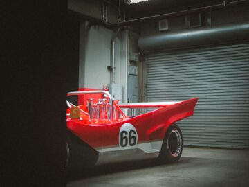 Een rood-witte raceauto, versierd met het nummer 66, staat in een slecht verlichte garage voor een gesloten metalen roldeur. Dit opvallende voertuig is niemand minder dan de legendarische Lotus Type 66.