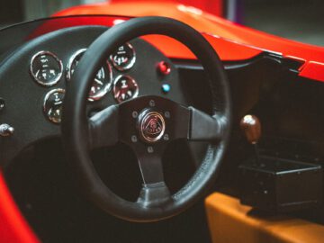 Close-up van het dashboard en het stuur van een auto met meters en controle-indicatoren, met het Lotus-embleem op het stuur, dat doet denken aan het iconische Lotus Type 66-ontwerp.