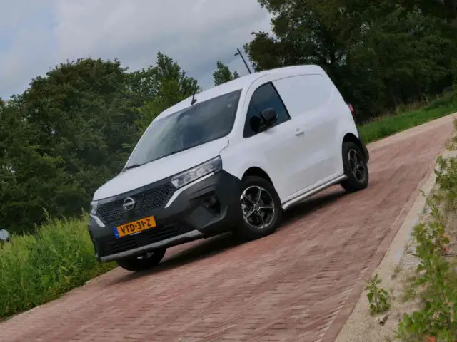 Een witte Nissan Townstar NV250-bestelwagen staat geparkeerd op een geplaveide oprit, omgeven door bomen en groen.