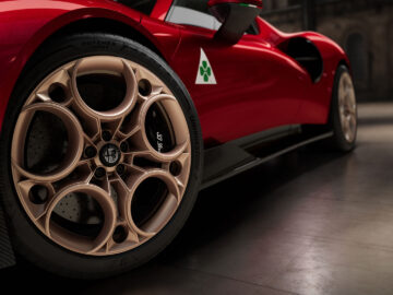 Close-up van de achterband en velg van een rode sportwagen, met een gedetailleerd ontwerp met cirkelvormige patronen. De auto, een Alfa Romeo 33 Stradale, heeft een wit embleem met een groen klaverblad op de zijkant.