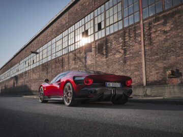 Op straat staat een strakke, rode Alfa Romeo 33 Stradale geparkeerd voor een groot bakstenen gebouw met veel ramen en een verweerde uitstraling. De auto heeft opvallende achterlichten en een kentekenplaat met de tekst "AR-033-ST.