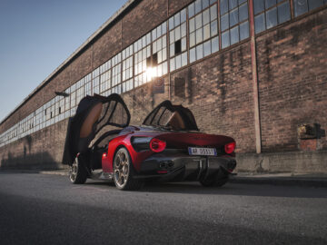 Rode Alfa Romeo 33 Stradale met open vleugeldeuren, geparkeerd in een lege straat naast een bakstenen gebouw met grote ramen. Op het kenteken staat "AR 0353T.