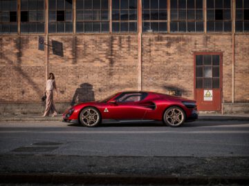 Een rode Alfa Romeo 33 Stradale staat op straat geparkeerd voor een industrieel bakstenen gebouw. Een persoon die lichtgekleurde kleding draagt, loopt op het trottoir vlakbij.