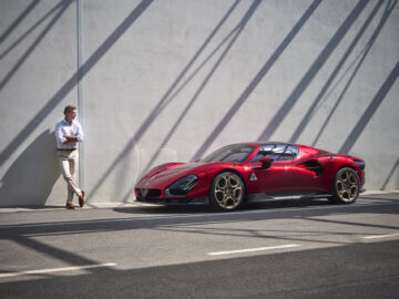 Een man in een wit overhemd en een beige broek staat tegen een muur, zijn blik gericht op een Alfa Romeo 33 Stradale met gouden velgen die op straat geparkeerd staat. Schaduwen van balken creëren patronen op de muur en de grond.