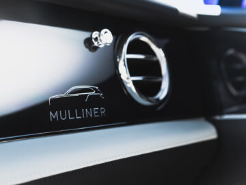 Close-up van een Bentley Bentayga EWB Mulliner-interieur met een gepolijste verchroomde ventilatieopening en het "Mulliner"-logo op een glanzend zwart paneel.