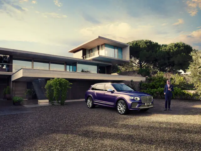 Een paarse Bentley Bentayga EWB Mulliner luxe SUV staat geparkeerd voor een modern huis. Rechts staat een persoon naar het voertuig te kijken. Het huis beschikt over grote ramen, een balkon en het omliggende groen.