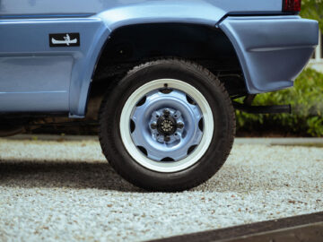 Close-up van het achterwiel van een blauwe Fiat Panda 4x4 Piccolo Lusso met een witte velg, geparkeerd op een grindoppervlak.