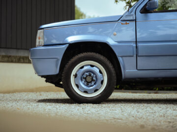 Close-up van de voorkant en het stuur van een blauwe Fiat Panda 4x4 Piccolo Lusso, geparkeerd op een grindoppervlak met een deel van een gebouw op de achtergrond.