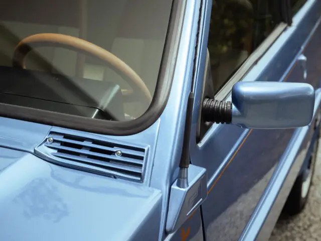 Close-up van de zijspiegel en voorruit van een blauwe vintage auto, waarbij een deel van het houten stuur door het glas zichtbaar is: een gedetailleerde blik op de charmante Fiat Panda 4x4 Piccolo Lusso.