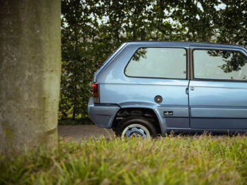 Zijaanzicht van een blauwe Fiat Panda 4x4 Piccolo Lusso geparkeerd in een straat vlakbij een boom, met groen gebladerte op de achtergrond.