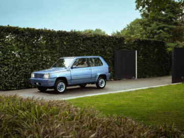 Een blauwe Fiat Panda 4x4 Piccolo Lusso staat geparkeerd op een oprit naast een hoge groene heg. Het tafereel toont een goed onderhouden tuin en een gedeeltelijk zichtbare poort. De lucht is helder.