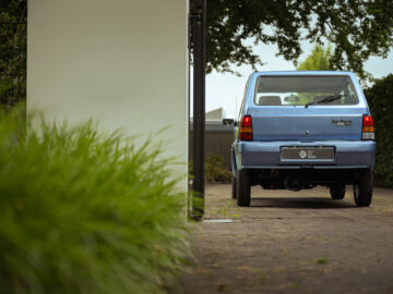 Een lichtblauwe Fiat Panda 4x4 Piccolo Lusso staat geparkeerd op een bakstenen oprit naast een witte muur, met groen gebladerte op de voorgrond en bomen op de achtergrond.