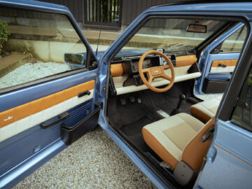 Een klassieke Fiat Panda 4x4 Piccolo Lusso met een blauw exterieur en beige interieur. Zowel de bestuurders- als de passagiersdeur staan open, waardoor een vintage stuurwiel en een eenvoudig dashboardontwerp zichtbaar zijn. De auto staat geparkeerd op een grindoppervlak en straalt een tijdloze charme uit.