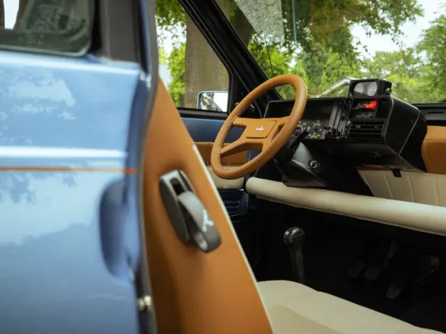 Interieur van een vintage Fiat Panda 4x4 Piccolo Lusso, met een blauwe deur, bruine accenten en het stuur. Het tafereel wordt gedeeltelijk aan het zicht onttrokken door een open raam, waardoor buiten bomen en groen zichtbaar zijn.