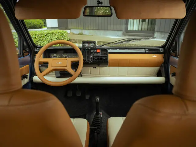 Interieur van een Fiat Panda 4x4 Piccolo Lusso met een bruin stuur, lichtbruine stoelen, handmatige versnellingspook en dashboard met verschillende bedieningselementen en meters. Door de voorruit is zicht op het buitengroen zichtbaar.