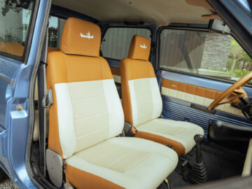 De afbeelding toont het interieur van een vintage Fiat Panda 4x4 Piccolo Lusso met beige en bruin lederen stoelen, een blauw deurpaneel en een bruin stuur. De hoofdsteunen zijn voorzien van een klein vliegtuiglogo.