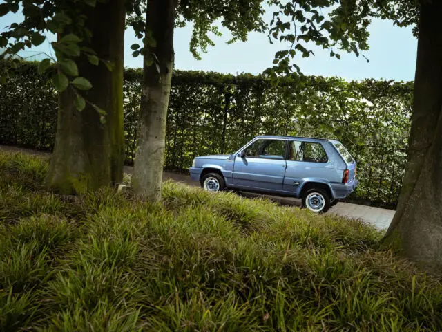 Een blauwe Fiat Panda 4x4 Piccolo Lusso staat geparkeerd op een pad naast een hoge, lommerrijke heg en bomen.