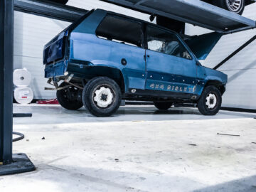 Een blauwe Fiat Panda 4x4 Piccolo Lusso-auto wordt op een hydraulisch platform in een garage gehesen, met de voorkap open. De garagevloer is schoon en op de achtergrond zijn diverse gereedschappen en benodigdheden zichtbaar.