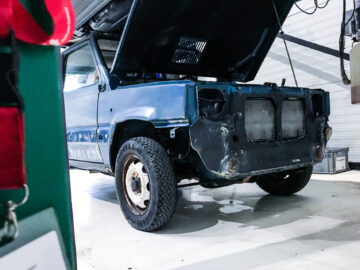 Een blauwe Fiat Panda 4x4 Piccolo Lusso met verhoogde motorkap staat in een garage en mist de grille en koplampen. De identiteitsbadge en het rode koord van een persoon zijn gedeeltelijk zichtbaar op de voorgrond.