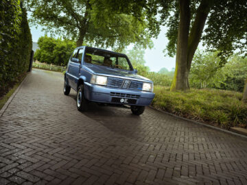 Een blauwe Fiat Panda 4x4 Piccolo Lusso staat geparkeerd op een bakstenen oprit met visgraatmotief, omgeven door weelderig groen en hoge bomen.