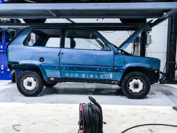 Een blauw Fiat Panda 4x4 Sisley-voertuig wordt in een garage getakeld met de motorkap open voor onderhoud. Het onderste deel van een uitschuifbare trap en enkele mechanische uitrusting bevinden zich op de voorgrond, waar de Piccolo Lusso te zien is.