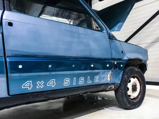 Een blauw Fiat Panda 4x4 Sisley-voertuig met zichtbare roest en afgebladderde verf aan de zijkant, binnen geparkeerd met de deur en de motorkap open.