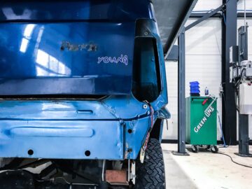 Een gedeeltelijk gedemonteerd blauw busje met zichtbare bedrading staat in een garage en laat de linkerachterzijde zien. Op het busje staat het woord ‘jong’ geschreven. Op de achtergrond zijn gereedschappen en een groene machine met het opschrift "GREENLINE" te zien, waardoor het tafereel lijkt op een reparatiecentrum van een Fiat Panda 4x4 Piccolo Lusso.