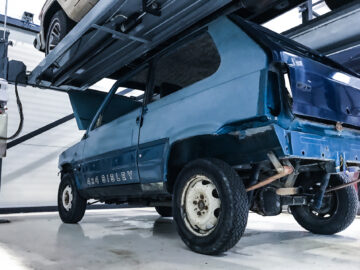 Een blauwe Fiat Panda 4x4 Piccolo Lusso staat op een hydraulische lift in een garage, waardoor het onderstel en de achterbanden zichtbaar zijn.