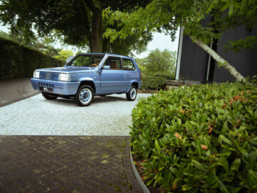 Een blauwe, vintage Fiat Panda 4x4 Piccolo Lusso compacte SUV staat geparkeerd op een onverharde oprit naast een gebouw, omgeven door groen op een bewolkte dag.