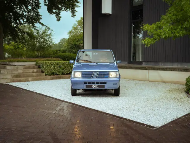 Een blauwe Fiat Panda 4x4 staat geparkeerd op een grindoprit naast een modern gebouw met grote ramen, omgeven door weelderig groen.