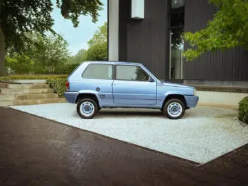 Een lichtblauwe Fiat Panda 4x4 Piccolo Lusso staat geparkeerd op een grindterrein voor een donkergrijs gebouw, omgeven door bomen en groen.