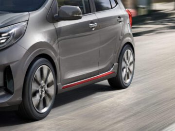 Close-up van een grijze compacte auto met lichtmetalen velgen die op een weg rijdt. De deur aan de bestuurderszijde en de voorpassagiersdeur zijn zichtbaar, wat het strakke ontwerp van een van de populairste auto's in Nederland benadrukt. De achtergrond toont een bewegingsonscherpte die de snelheid aangeeft.