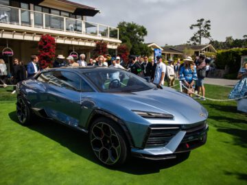 Op een groen gazon staat een strakke, blauwe luxe SUV, de Lamborghini Lanzador, met daarachter een menigte mensen. Vlakbij zijn een gebouw en bomen zichtbaar onder een gedeeltelijk bewolkte hemel.