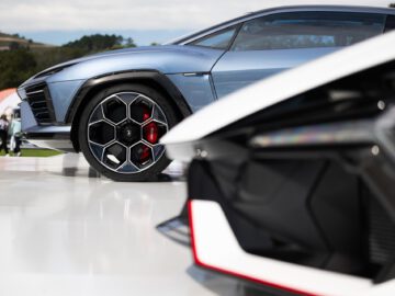 Close-up van de wielen en gedeeltelijke carrosseriedelen van twee luxe sportwagens, waaronder een Lamborghini Lanzador, met opvallende geometrische wielontwerpen en strakke, aerodynamische structuren, buiten tentoongesteld op een reflecterend oppervlak.