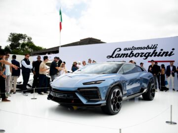 Een blauwe Lamborghini Lanzador SUV wordt buiten op een wit platform tentoongesteld, omringd door mensen. Op de achtergrond is het Lamborghini-logo en de branding te zien. Op de achtergrond is een Italiaanse vlag zichtbaar.