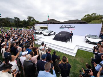 Een grote menigte verzamelt zich rond een podium met drie Lamborghini-auto's, waarvan één overdekt, op een buitenevenement met een luidspreker onder een tent, reikhalzend uitkijkend naar de onthulling van de nieuwe Lamborghini Lanzador.