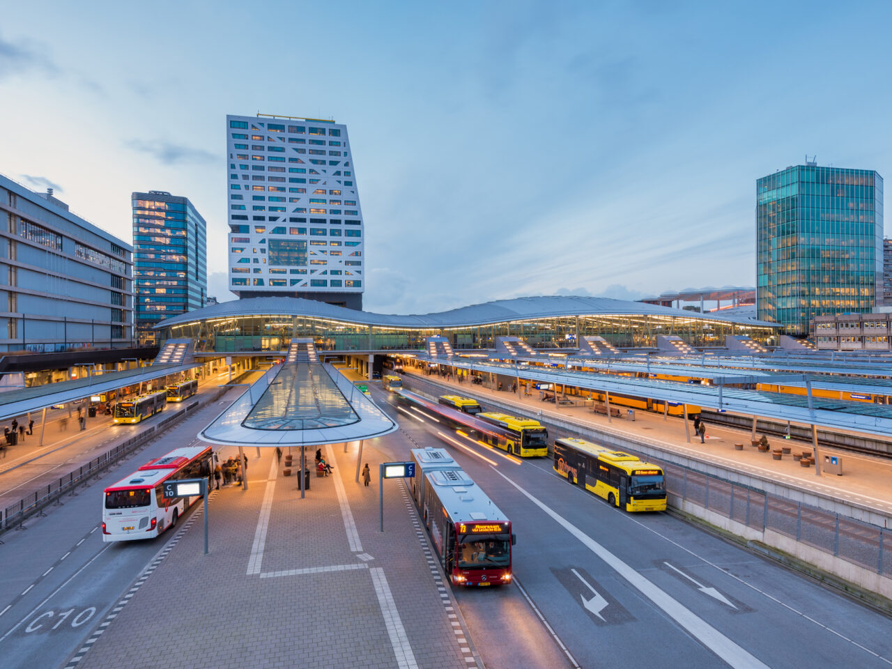 Een modern busstation met meerdere Nederland OV-bussen geparkeerd op verschillende perrons, omringd door hoge gebouwen in de schemering.
