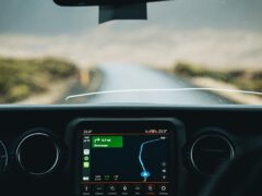 Uitzicht vanuit een auto met het Apple CarPlay-navigatiesysteem op het dashboard, dat een route aangeeft. Voor ons strekt zich een natte, lege weg uit, zichtbaar door de voorruit.