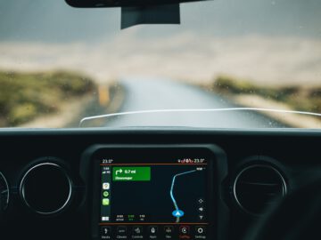 Uitzicht vanuit een auto met het Apple CarPlay-navigatiesysteem op het dashboard, dat een route aangeeft. Voor ons strekt zich een natte, lege weg uit, zichtbaar door de voorruit.
