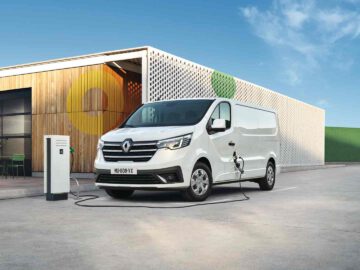 Een witte elektrische bestelwagen van Renault Trafic staat geparkeerd bij een laadstation buiten een modern houten gebouw met geometrische muurontwerpen. Het busje is aangesloten op de oplader, terwijl de lucht erboven helder is met wat wolken.