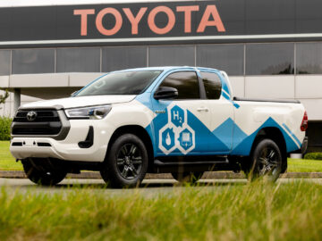 Een wit-blauwe Hilux-pick-up met waterstofbrandstofcelafbeeldingen staat geparkeerd voor een Toyota-gebouw.