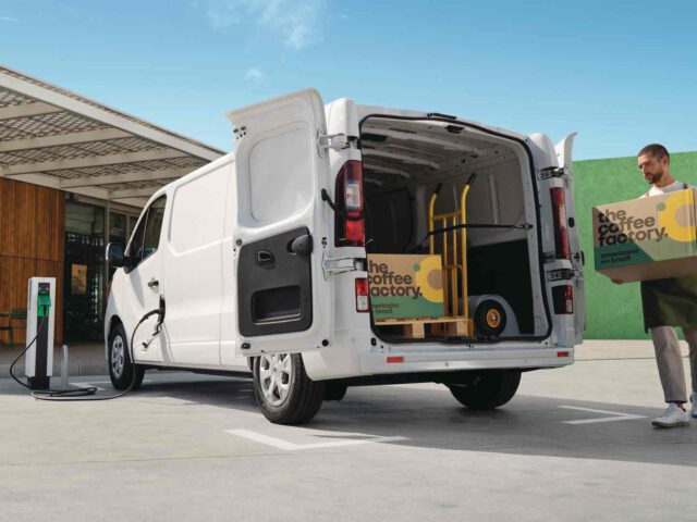 Een man draagt een doos met het opschrift 'The Coffee Factory' naar een open witte bestelwagen van Renault Trafic, geparkeerd bij een laadstation voor elektrische voertuigen.