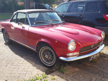 Een rode vintage Fiat 124 Sport Spider cabriolet staat geparkeerd in een zonovergoten straat naast een zwarte SUV.