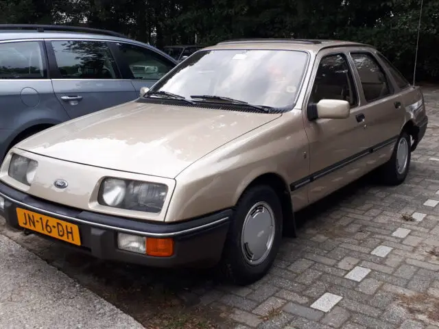 Op een parkeerplaats staat een beige Ford Sierra sedan met links een grijze auto. De auto heeft een Nederlands kenteken met de code JN-16-DH. Op de achtergrond zijn bomen zichtbaar.