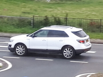 Een witte Saab 9-4X SUV rijdt op een weg in de buurt van een grasveld en navigeert door een gebogen gedeelte met zwart-witte wegmarkeringen. Op de achtergrond zijn een hek en een veld zichtbaar.