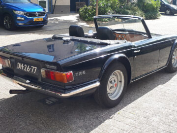 Een marineblauwe Triumph TR6-cabriolet geparkeerd in een zonovergoten straat met de kap naar beneden en trots een kentekenplaat DH-26-77 tonend.
