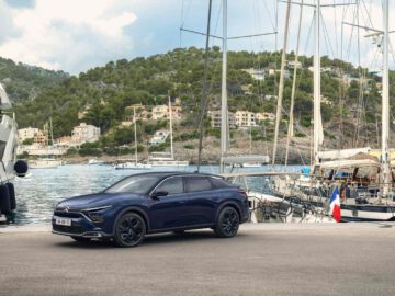 Een donkerblauwe C5 X SUV staat geparkeerd aan de waterkant, met zeilboten en heuvels vol gebouwen op de achtergrond.