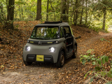 Een compacte, grijze Citroën Ami Buggy rijdt op een met bladeren bedekt bospad, omgeven door bomen met groen en bruin blad.