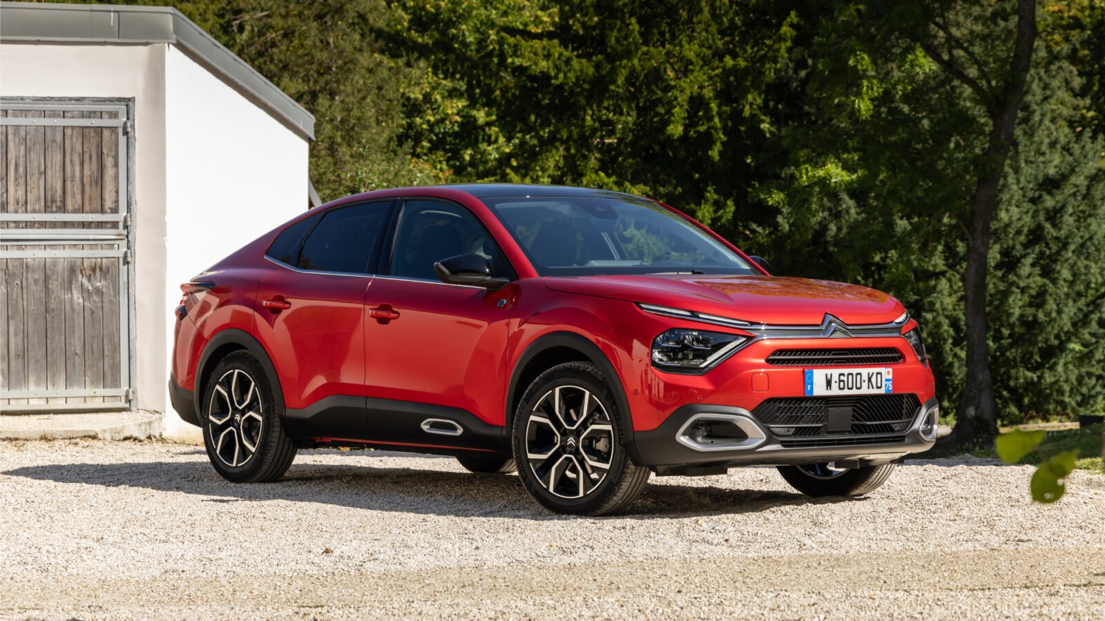 Een rode Citroën e-C4 X SUV staat geparkeerd op een grindoppervlak buiten een wit gebouw met houten deuren.