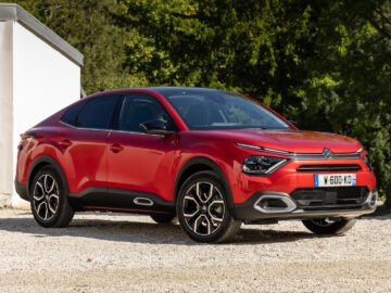 Een rode Citroën e-C4 X SUV staat geparkeerd op een grindoppervlak buiten een wit gebouw met houten deuren.